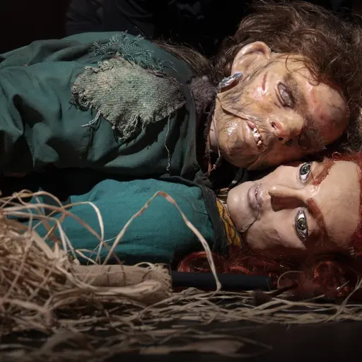 Två dockor ligger sida vid sida på marken, omgivna av halm. Den ena har mörkt hår och skadade kläder medan den andra har rött hår och en neutral ansiktsuttryck. Mörk bakgrund med svaga färger.