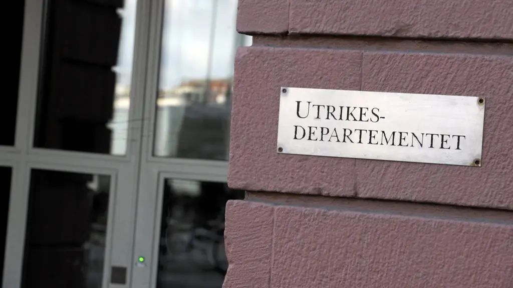 En närbild av en skylt med texten 'Utrikesdepartementet' monterad på en tegelvägg. I bakgrunden syns glasdörrar och en suddig vy av stadsmiljö.