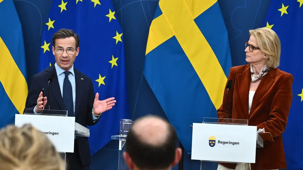 Statsministern och finansministern står vid varsin talarstol under en pressträff i Stockholm. Bakgrunden pryds av svenska och EU-flaggor. Flera personer i publiken syns i förgrunden.