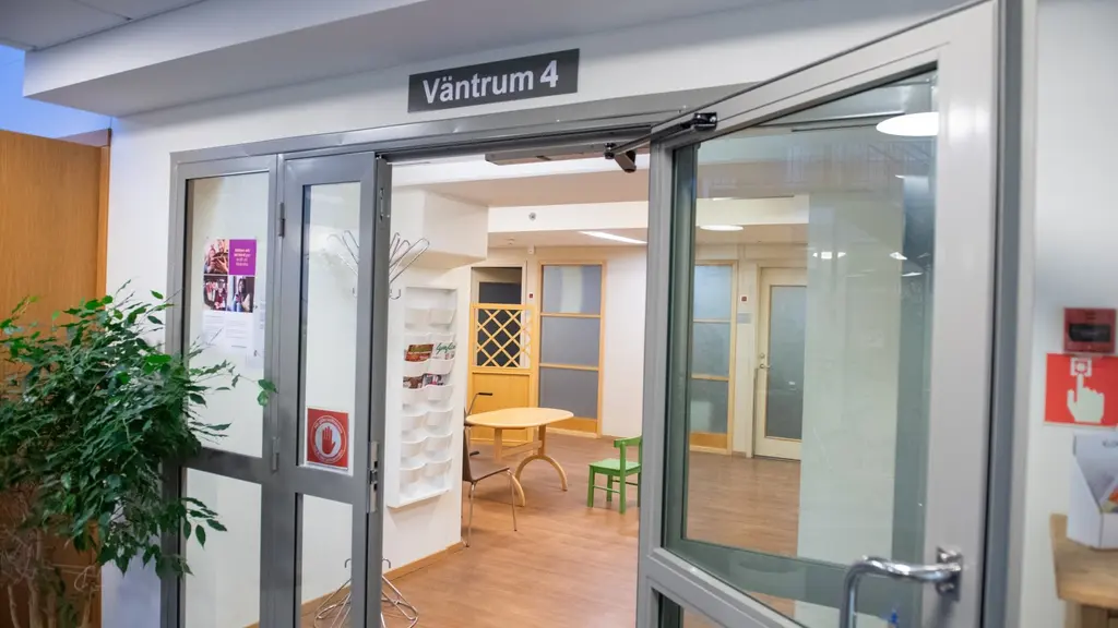 Ingång till ett väntrum på en vårdcentral med skylt 'Väntrum 4'. Glasdörrar leder in till ett ljust rum med stolar och bord. En grön växt står vid sidan av dörren. Brandsläckare syns på väggen.