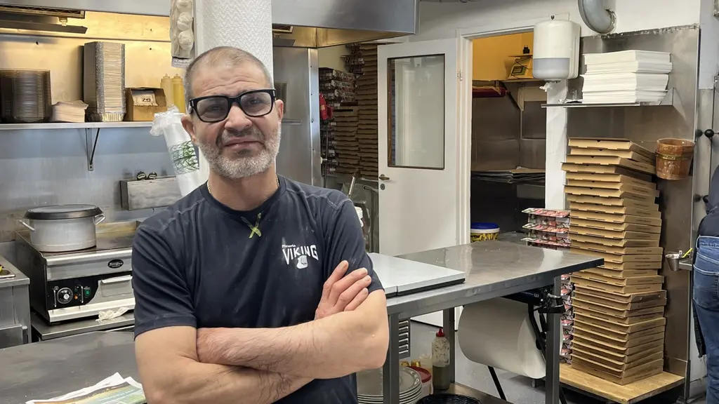 En man står med armarna i kors i ett kök med professionell utrustning. Bakom honom syns staplade pizzakartonger, en ugn och köksredskap. Miljön tyder på att det är en pizzeria.