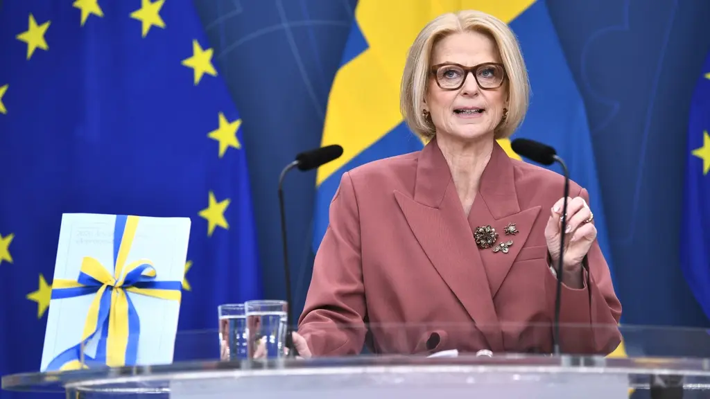 Finansminister Elisabeth Svantesson står vid ett podium och presenterar vårbudgeten 2026. Bakgrunden visar EU:s och Sveriges flaggor. En budgetrapport med blått och gult band ligger bredvid.