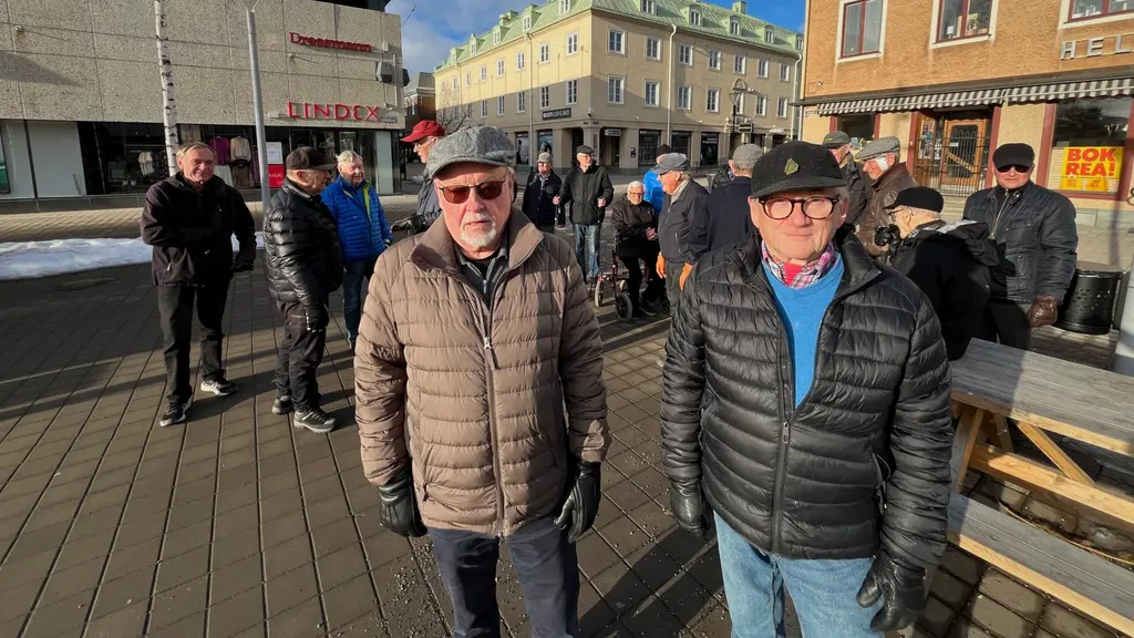 Två personer står i förgrunden på ett torg med vinterkläder. Bakom dem syns flera människor i en grupp, byggnader och en solig vintermiljö med snö på marken.
