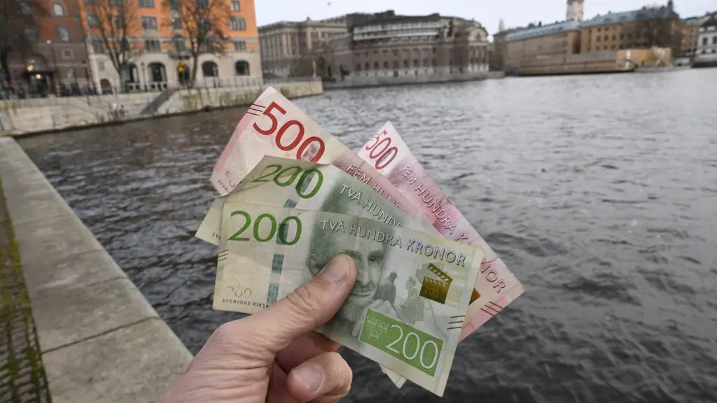 En hand håller upp svenska sedlar, inklusive 200- och 500-kronorssedlar, framför en vattenvy i centrala Stockholm med historiska byggnader i bakgrunden.