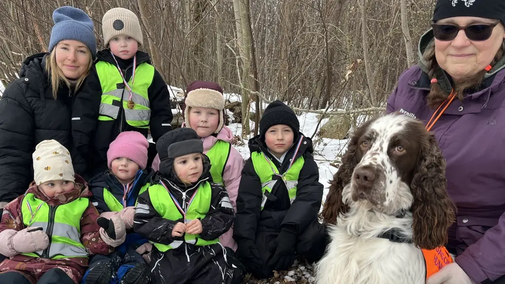 En grupp barn med reflexvästar sitter på marken tillsammans med en stor, vit och brun hund. En vuxen person håller hunden i ett orange koppel. Bakgrunden består av snö och träd i en utomhusmiljö.