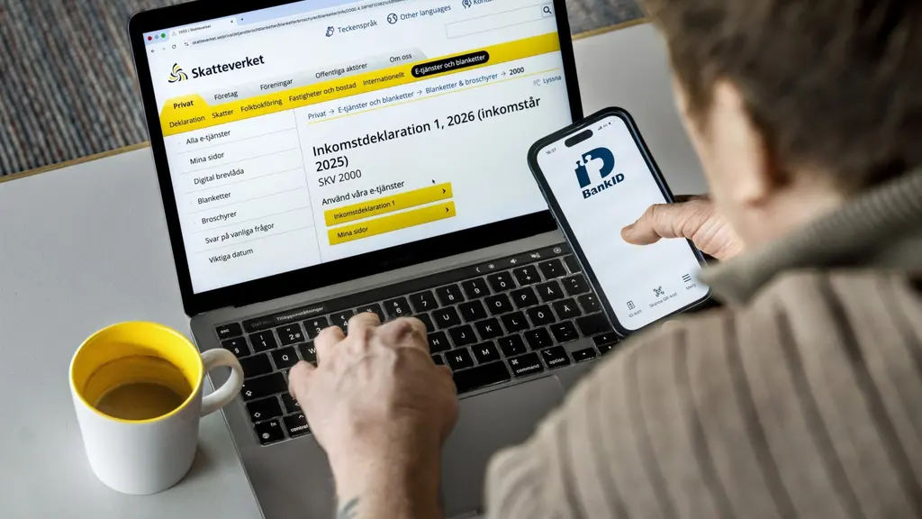 En person sitter vid ett bord och deklarerar digitalt på Skatteverkets webbplats via en bärbar dator. På mobilskärmen syns en app med BankID. En gul kaffekopp står bredvid datorn.