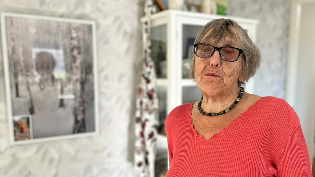 En äldre kvinna med kort hår står i ett rum med ljusa väggar och en bokhylla i bakgrunden. Hon bär en korallröd stickad tröja och ett halsband. På väggen hänger en tavla med naturmotiv.