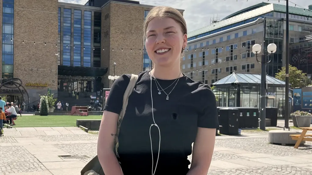 En ung kvinna iklädd jeans och t-shirt står på ett torg, det är fint väder och solen skiner. 