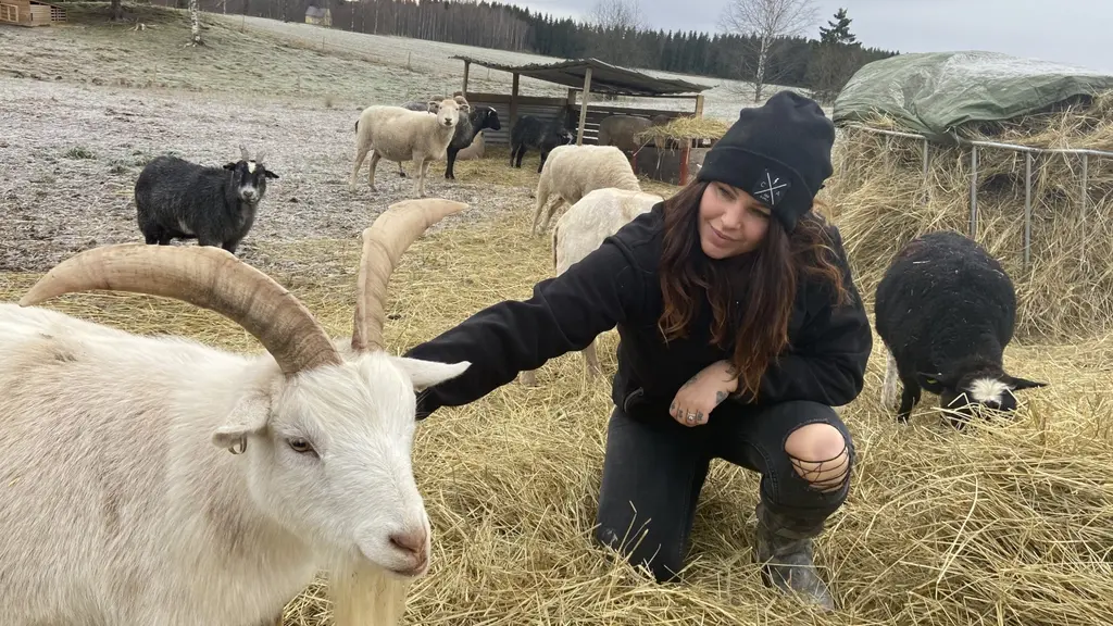 På fotot syns Cassandra. Hon har svarta kläder och en mössa på huvudet. Hennes hand är tatuerad. Hon klappar en get.