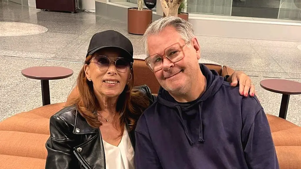 Sylvia Vrethammar syns till vänster på bilden. Hon bär en keps, tonade glasögon och skinnjacka som är uppknäppt. Hon håller vänster arm runt Micke Cederberg och man ser hennes hand på hans vänstra axel. Micke bär glasögon, är kortklippt och har en hoodtröja på sig.