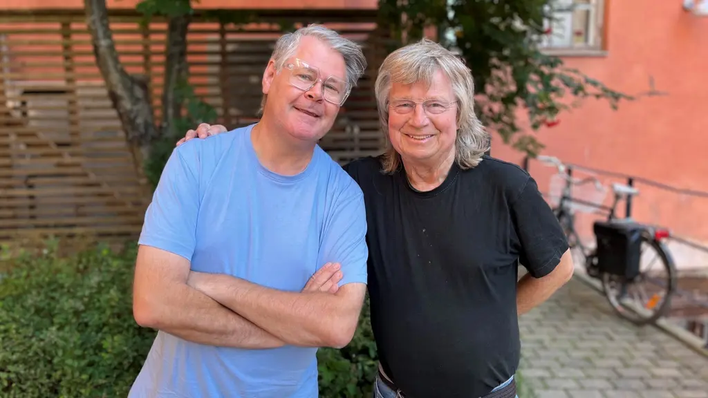 Micke Cederberg och Janne Schaffer står utomhus. Janne håller höger hand på Mickes vänstra axel. Båda bär glasögon. Man ser träd och buskar i bakgrunden samt en cykel.