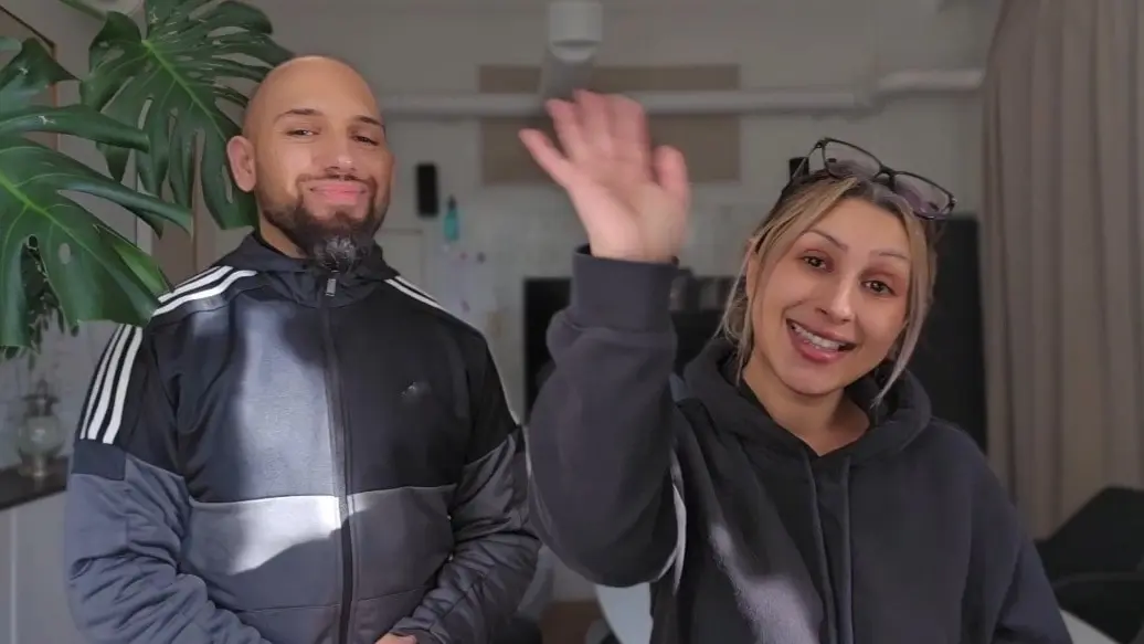 En man och kvinna tittar in i kameran. Kvinnan vinkar med ena handen. 