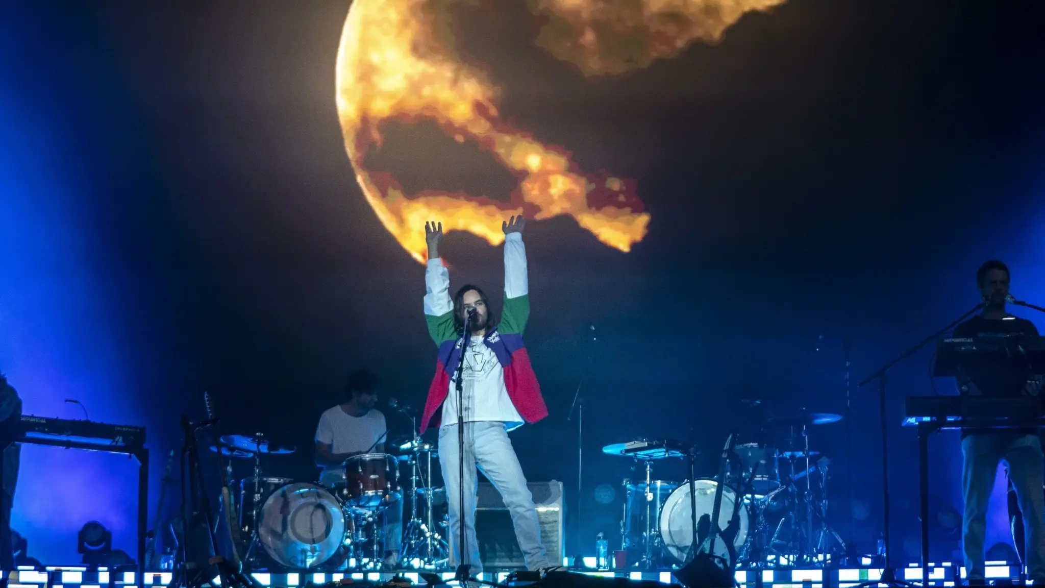 Bilden visar ett foto på ett liveframträdande av bandet Tame Impala på scen. Längst fram i mitten ser man frontfiguren och sångaren Kevin Parker och i bakgrunden syns två bandmedlemmar bakom varsitt trumset och två bandmedlemmar bakom varsin keybord. i Bakgrunden syns en stor måne projicerad. 