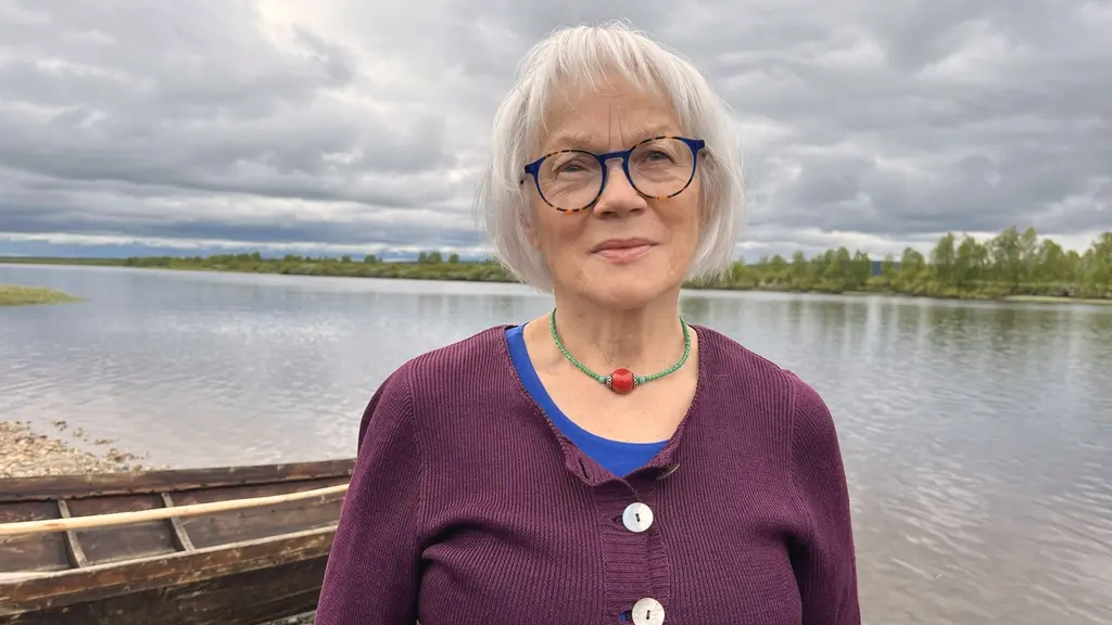 Porträttbild av Britta Marakatt-Labba, med vitt kort hår, glasögon med svarta skalmar. I bakgrunden rinner Lainioälven.