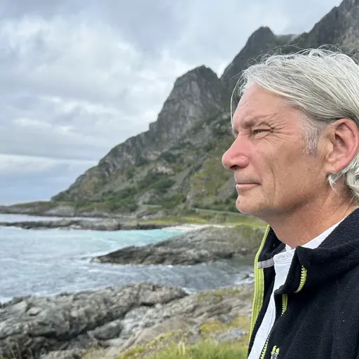 Halvbild i profil på Robert Vars-Gaup. I bakgrunden finns höga klippor och havet som slår in mot dem. Han är klädd i en mörk jacka med limegrönt blixtlås och har en vit skjorta under.