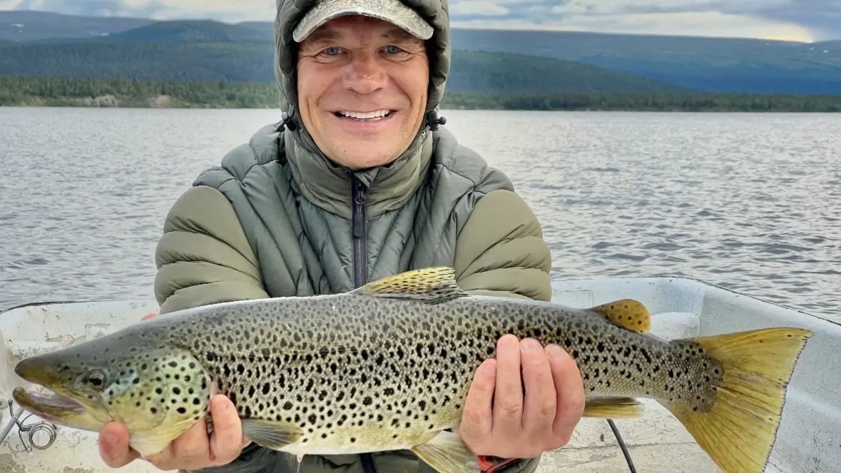 Man som sitter i en båt håller i en fisk och ler med fjäll i bakgrunden 