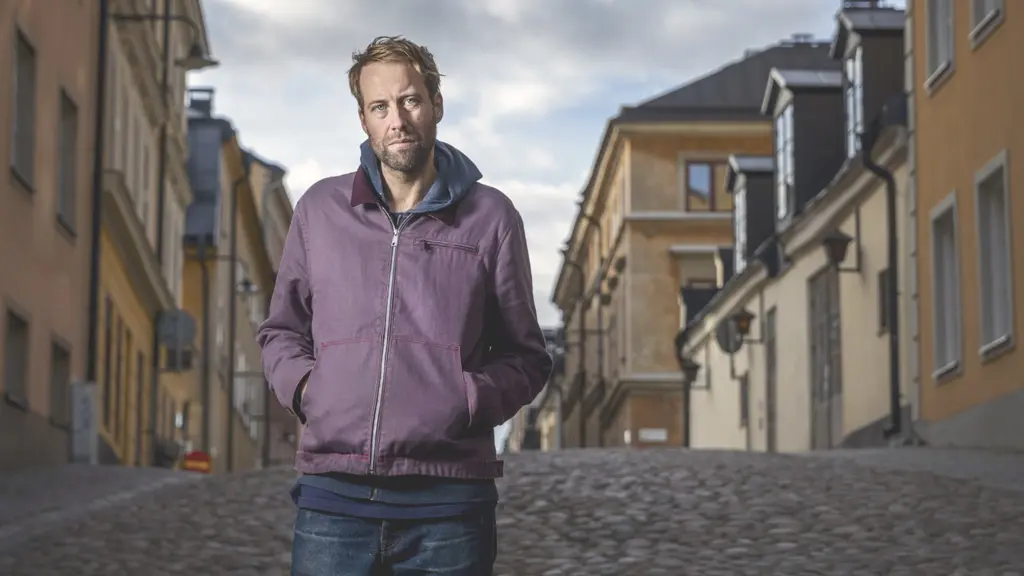 En man i lila jacka står på en gata med husfasader på sidorna.