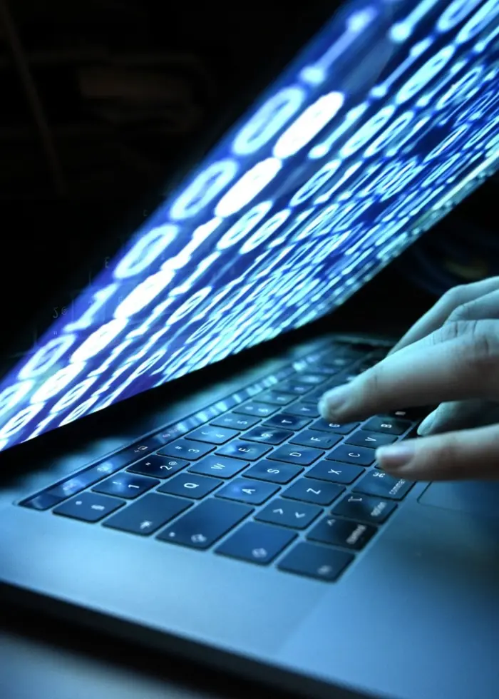 En närbild av en bärbar dator i mörker, där skärmen lyser starkt med ett digitalt mönster av blå kod. En hand skriver på tangentbordet, vilket illustrerar ämnet cybersäkerhet.