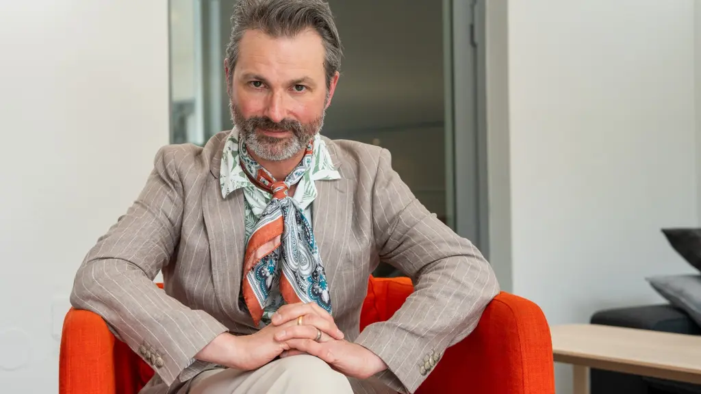 Ola Salo sitter i en röd fotölj. Han har på sig en randig kavaj och scarf. 