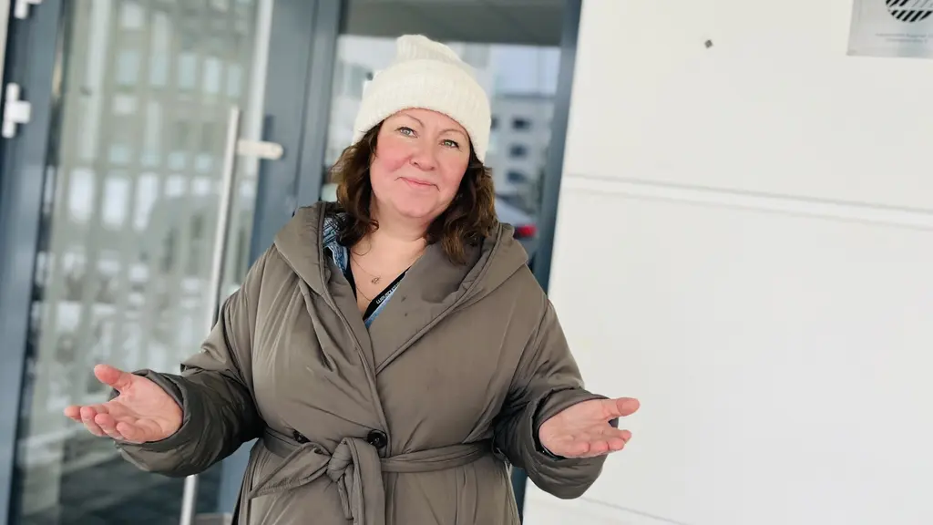  Victoria Herkules klädd i en brun jacka och vit mössa står utomhus framför en byggnad med glasdörrar. Hon har händerna uppsträckta i en gest som antyder diskussion eller förklaring.