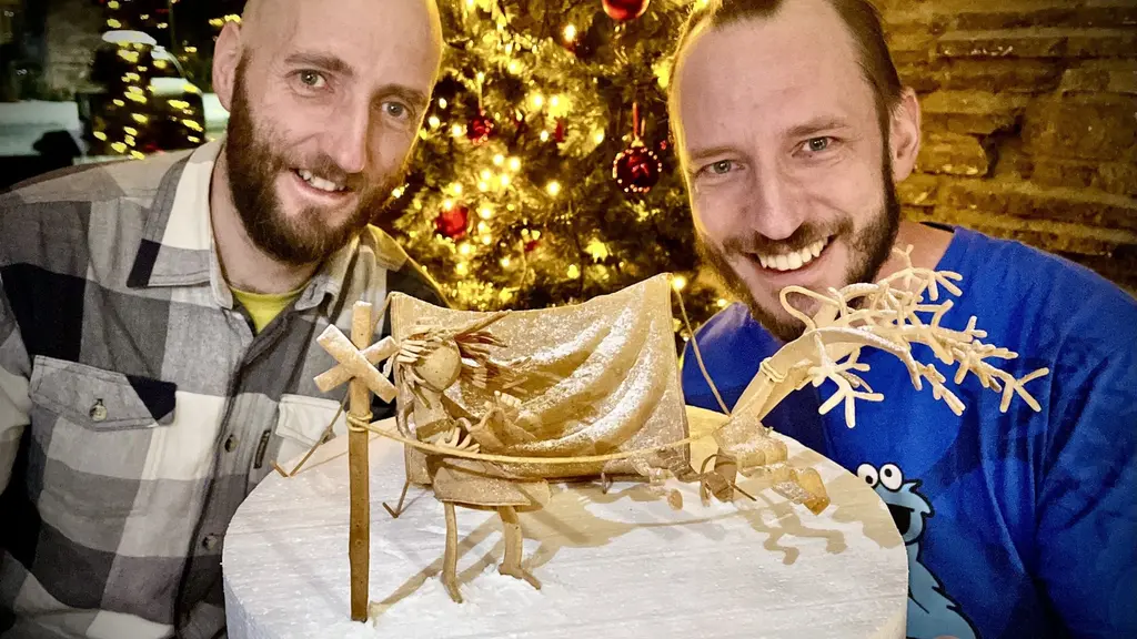 Ett detaljrikt pepparkaksbygge med temat vintercamping i snöstorm visas upp. Bakgrunden pryds av en julgran med guldbelysning och röda dekorationer. Bygget har tält och snöiga träd.