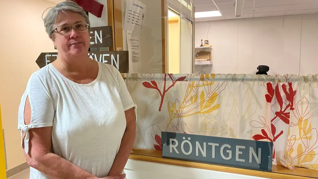 Kvinna i vit tröja och glasögon står vid en reception.Bredvid finns en skylt som det står "röntgen" på.