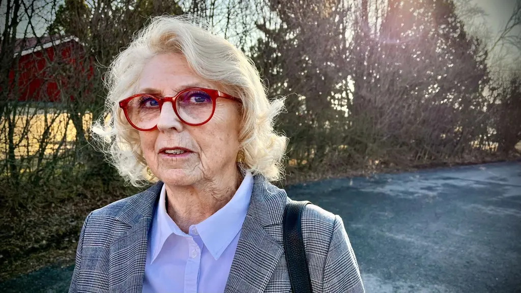 En kvinna i grårutig kavaj, ljus skjorta och röda glasögon. Hon står utomhus. 