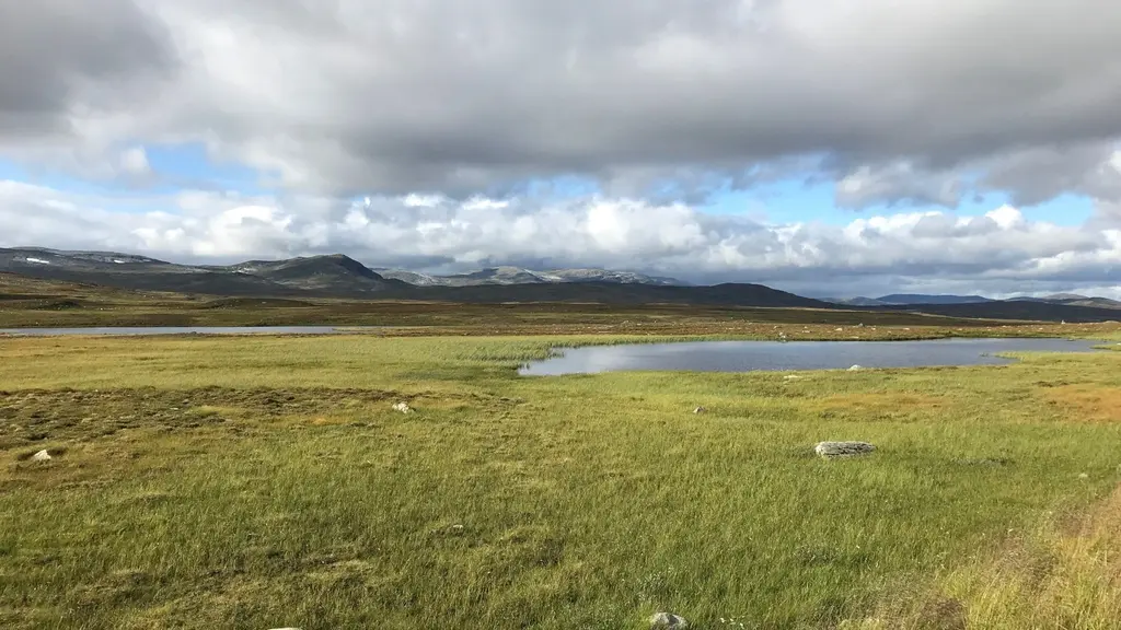 Vy över Stekenjokk med öppna fjällandskap, gröna ängar och en liten sjö i förgrunden. I bakgrunden syns berg med fläckar av snö under en himmel med moln och blåa inslag.