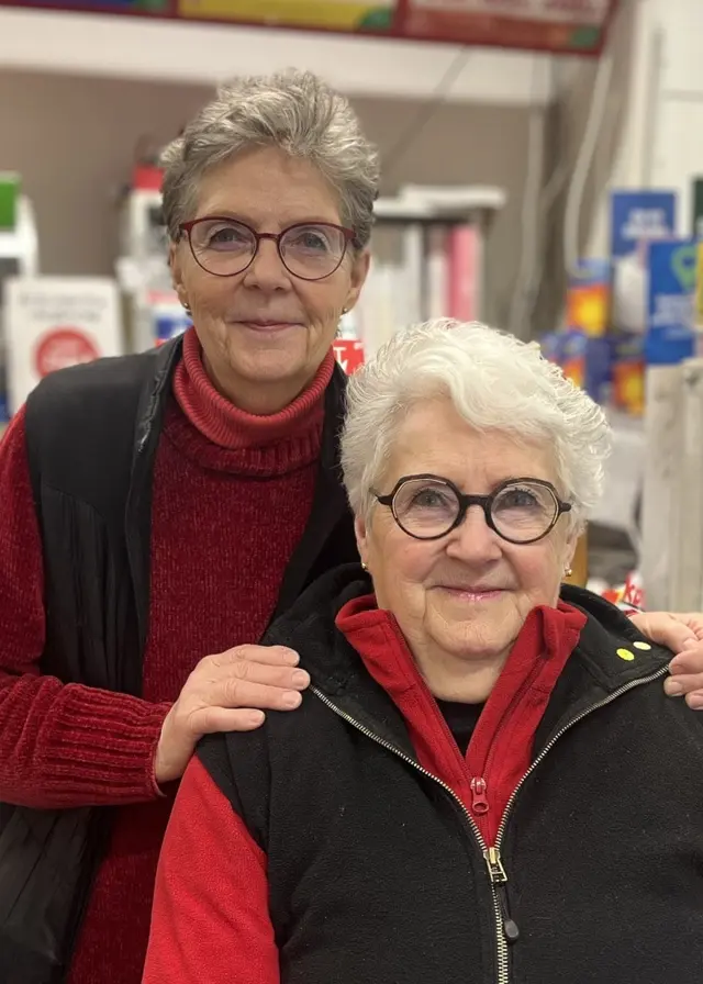 Två äldre kvinnor står bakom butikens kassadisk. De är klädda i röda tröjor och svarta västar. Hyllor med varor och skyltar syns i bakgrunden. De hoppas finna någon som kan ta över butiken i Sikås.