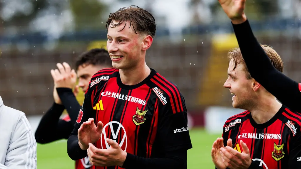 Philip Bonde i Östersunds FK:s röda och svarta matchtröja applåderar på fotbollsplanen, omgiven av lagkamrater. Bakgrunden visar läktare och regnigt väder.