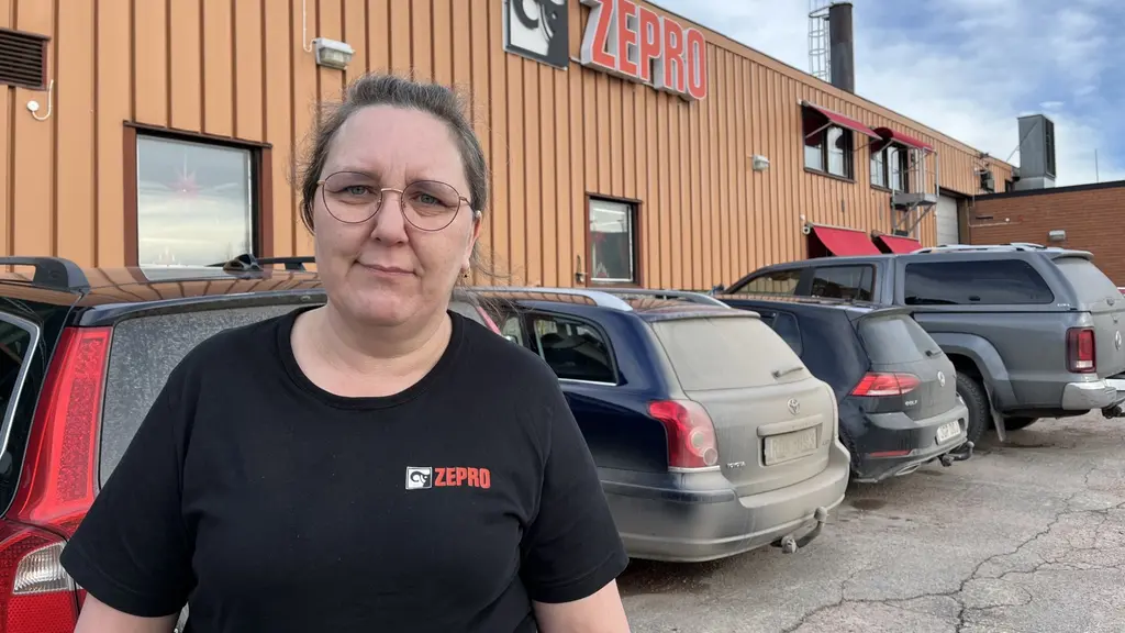 En person står framför en parkeringsplats med flera bilar och en byggnad i bakgrunden. Byggnaden har en skylt med texten 'Zepro' och stora fönster. Himlen är delvis molnig.