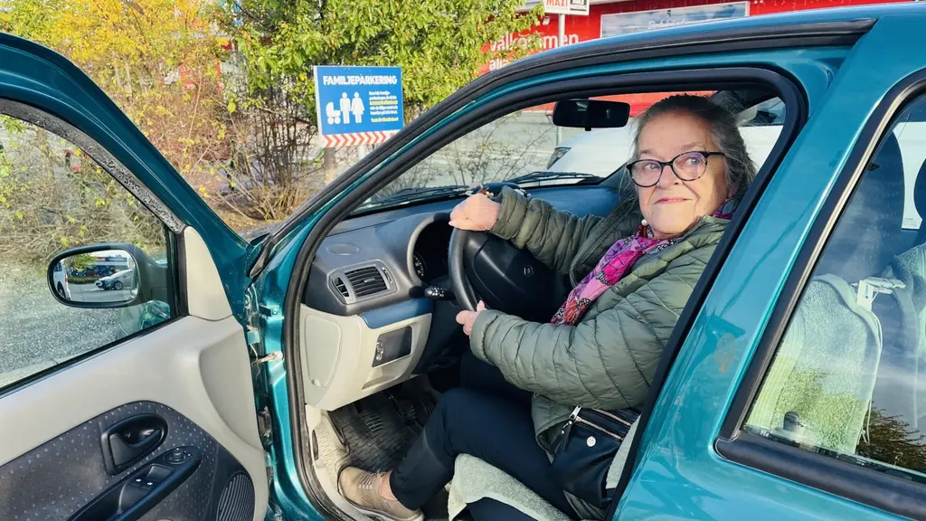 En person sitter i förarsätet på en grön bil som står parkerad utomhus vid en så kallad familjeparkering utanför en större butik. Bakgrunden visar träd och en byggnad med röda detaljer.