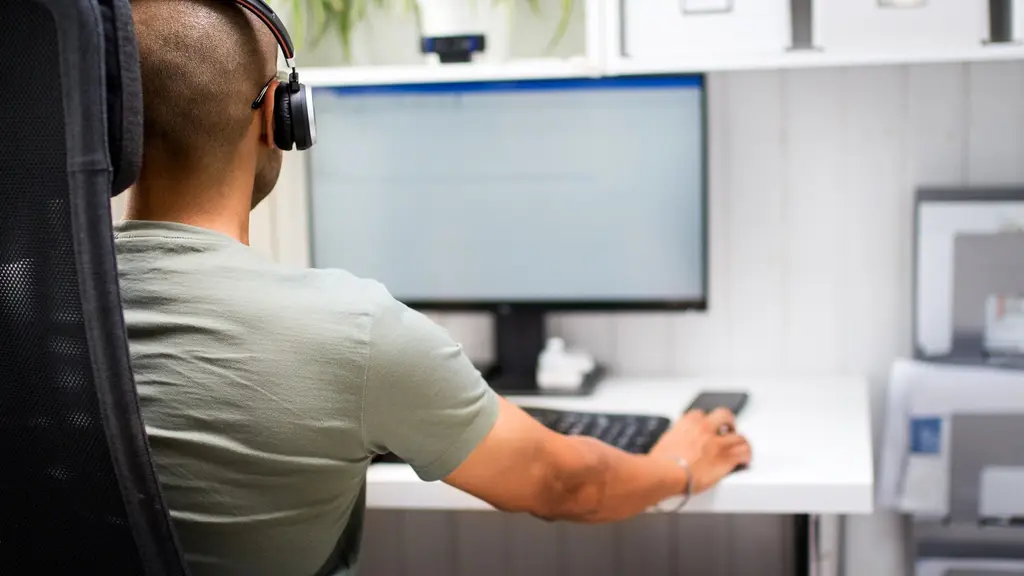 En person sitter i en hemmamiljö med hörlurar och arbetar vid en dator. På skrivbordet finns en mobiltelefon och kontorsutrustning