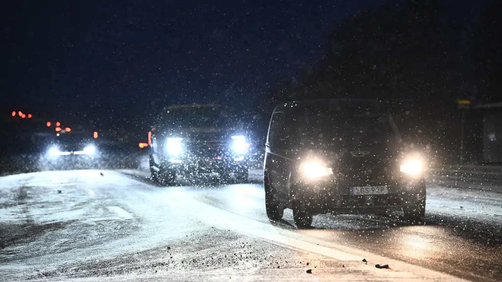 Bilar kör på en snöig riksväg 55 i Skolsta nära Enköping under kvällstid. Snöfall och mörker försvårar sikten, medan bilarnas strålkastare lyser upp den vinterklädda vägen.