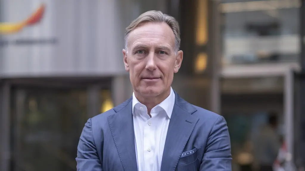 En man står framför en modern byggnad med glasfasad, klädd i en blå kavaj och vit skjorta. Bakgrunden visar ett kontorskomplex med en färgglad logotyp vid ingången.