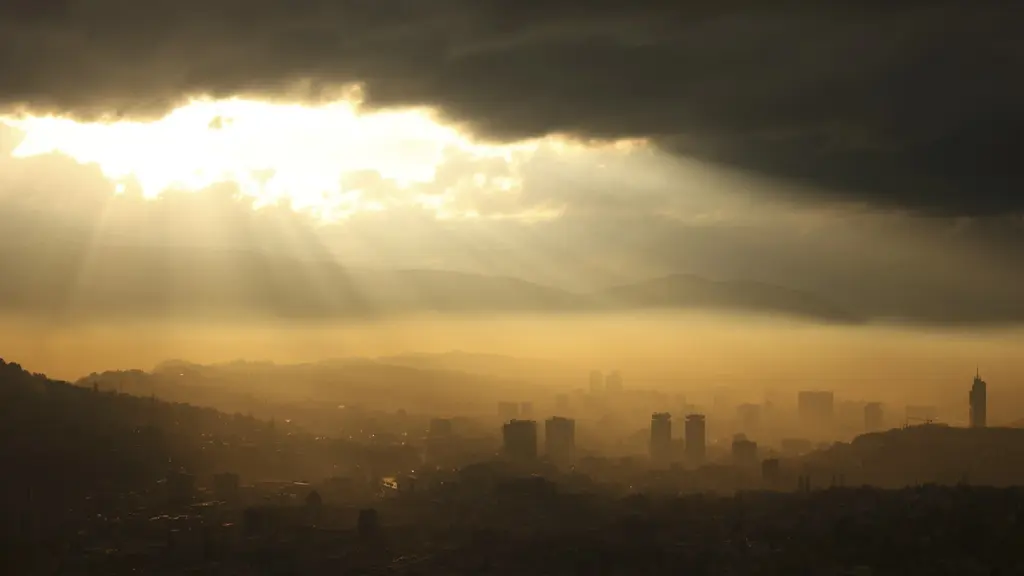Vy över Sarajevo vid solnedgång, täckt av smog. Solens strålar bryter igenom mörka moln och lyser svagt över staden. Byggnader och kullar syns i bakgrunden under det dimmiga landskapet.