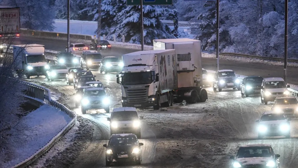 En lastbil med släp blockerar väg 73 mot Nynäshamn i Stockholm under kraftigt snöfall, vilket orsakar trafikproblem. Bilar står i kö på den snötäckta vägen omgivna av vinterlandskap.