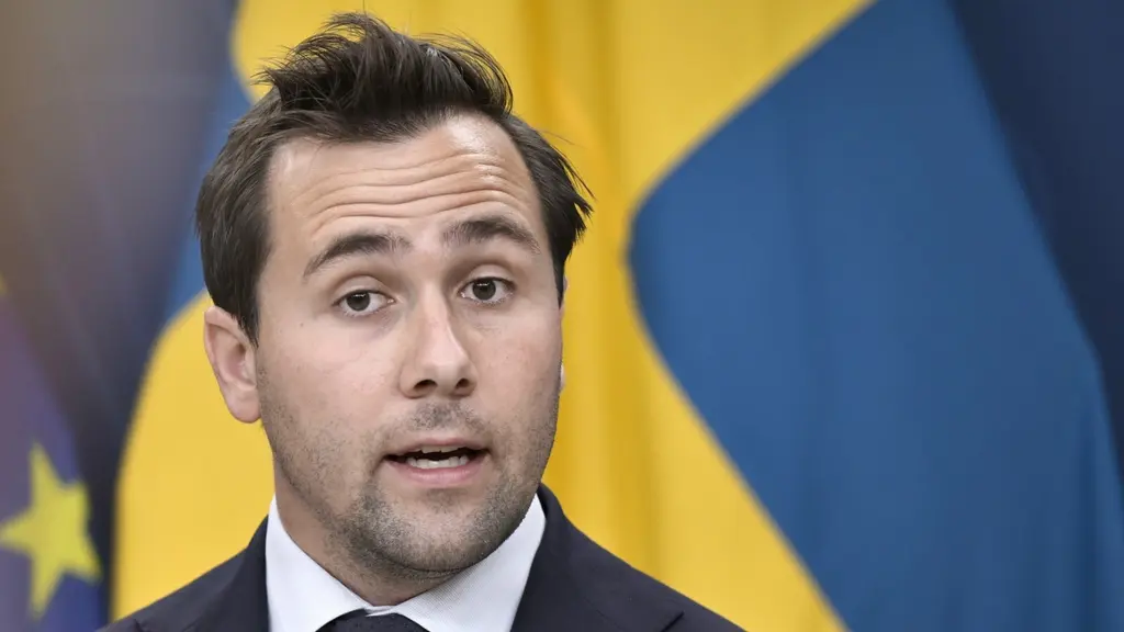 Benjamin Dousa, bistånds- och utrikeshandelsminister, står framför svenska och EU-flaggor under en pressträff om tullavtalet mellan EU och USA. Han bär kostym och slips.
