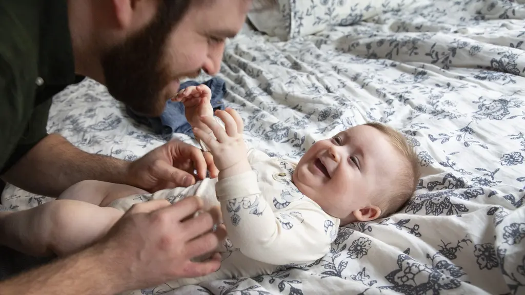 En pappa leker med sitt barn som ligger på en säng med blommigt överkast. Barnet är klädd i en ljus body och ser ut att skratta. Miljön är ljus och hemtrevlig.