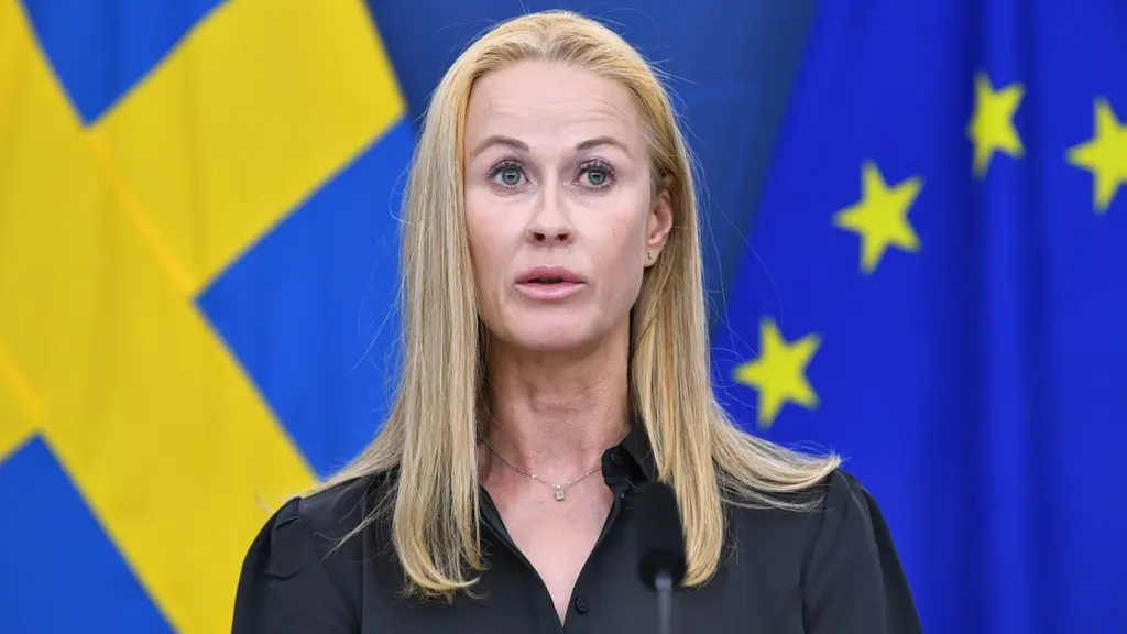 En kvinna med långt blont hår står framför svenska och EU-flaggor under en pressträff om nya åtgärder mot kriminaliteten. Hon är klädd i svart och talar vid en mikrofon.