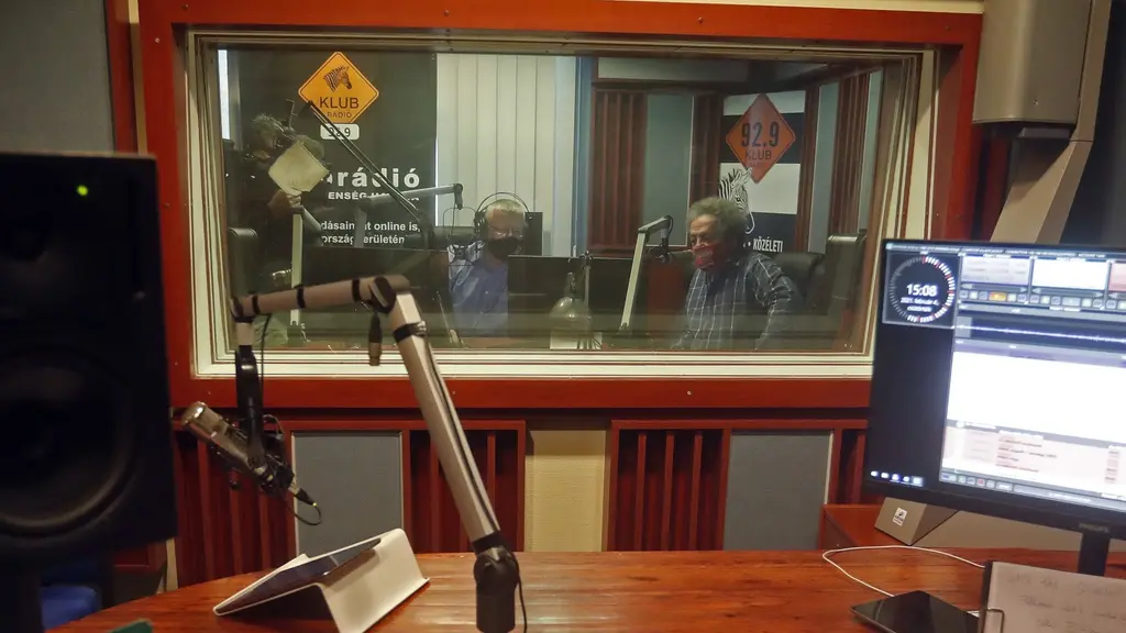 En radiostudio med mikrofoner, datorer och ljudutrustning syns i förgrunden. Bakom ett ljudisolerat glasfönster sitter två personer i samtal, med radioutrustning runtomkring.