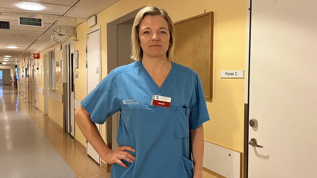 Allmänläkaren Lina Renström i ljusblå vårdkläder står i en sjukhuskorridor med gula väggar och stängda dörrar. 