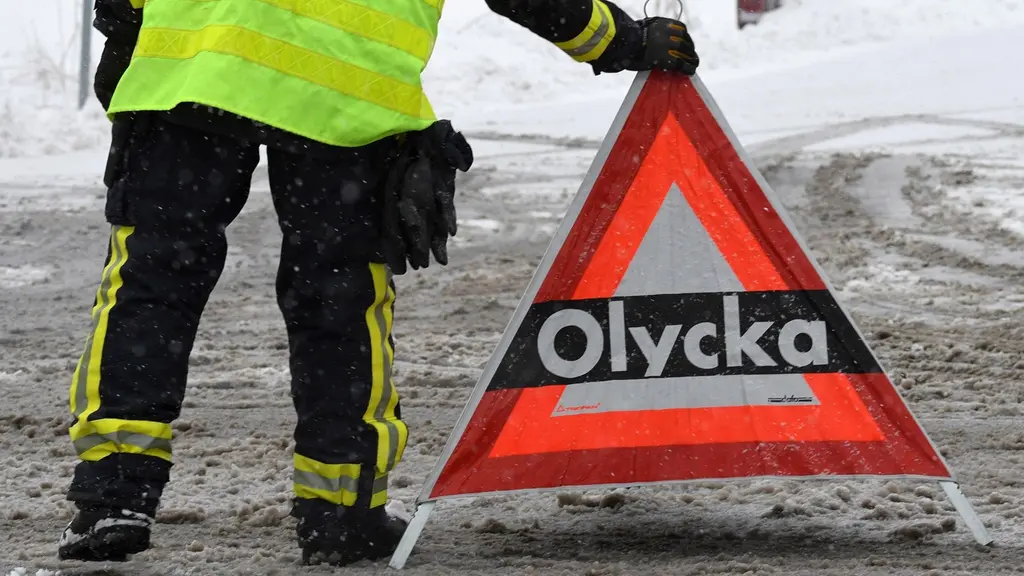 En person i reflexväst sätter ut en varningsskylt med texten "Olycka" på en snömoddig väg.