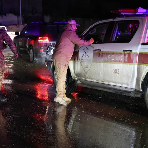 Syriska demokratiska styrkor (SDF) ses vid en väg med flera fordon, inklusive en militärpolisbil, på natten. Reflektioner av ljus från fordonen syns på den våta asfalten.