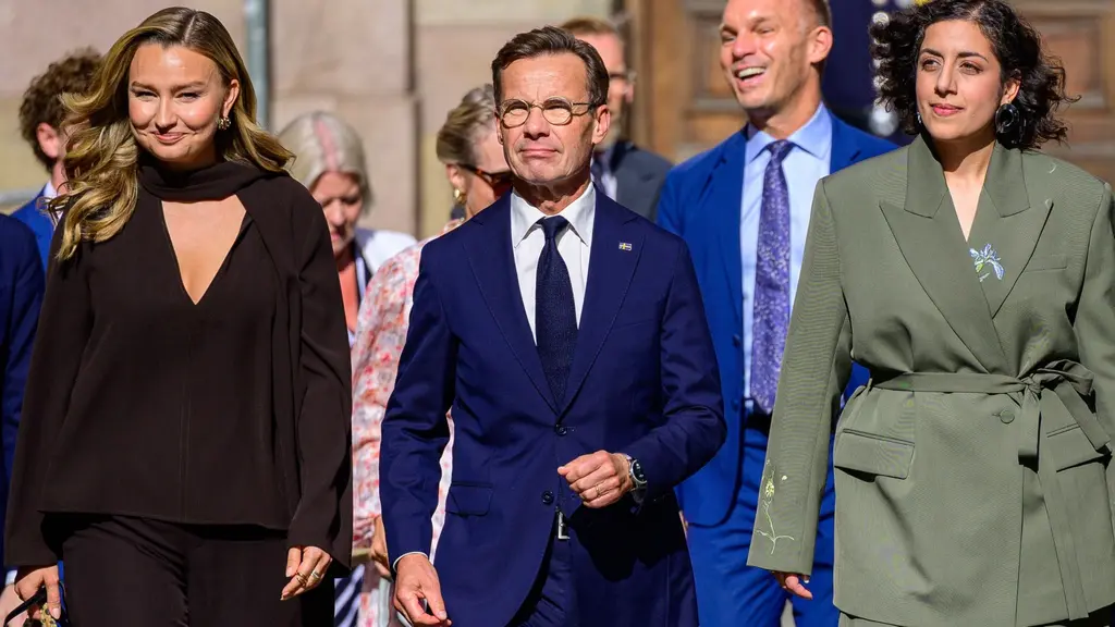 Tre personer i formella kläder promenerar mot kameran i solen. En man i mörkblå kostym går i mitten, flankerad av två kvinnor i mörka och ljusa outfits. Bakgrunden visar en historisk byggnad.