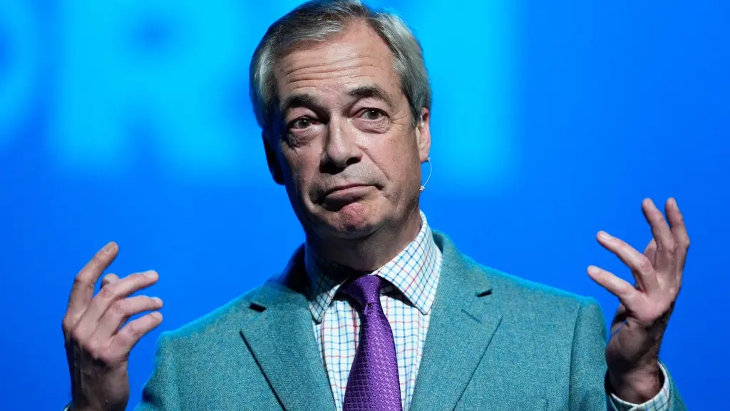Reform Partys ledare Nigel Farage talar på ett politiskt möte i Llandudno, Wales, med blå bakgrund och händerna höjda i en gest för att understryka sitt budskap.