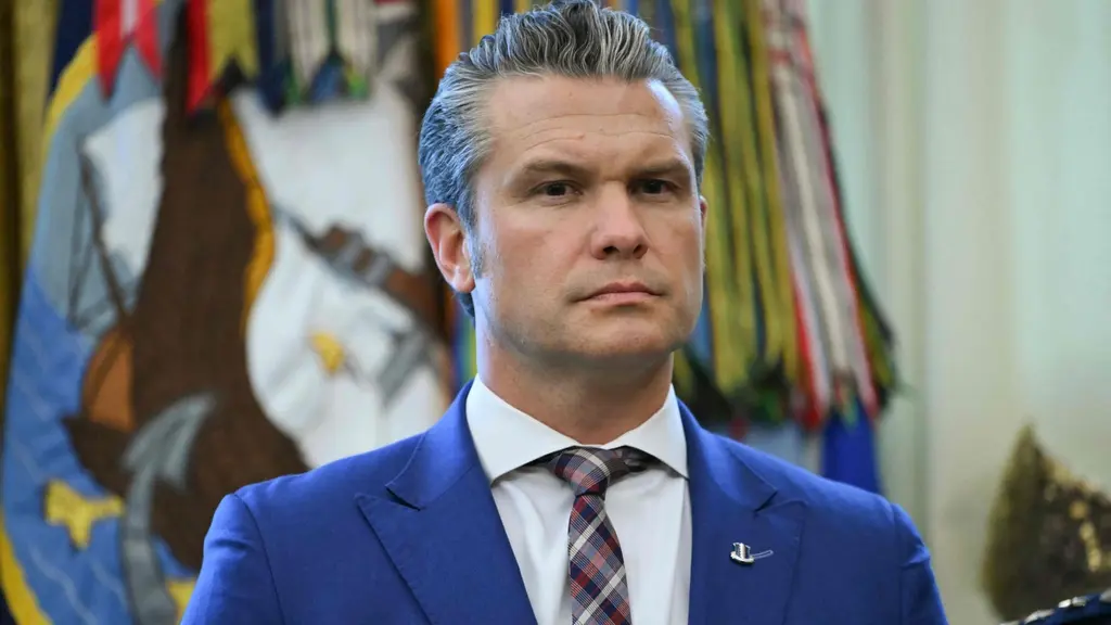 En person i kostym står framför en färgglad vapensköld och flaggor i en officiell miljö, möjligen på en plats som Vita huset. Personen verkar delta i en ceremoni eller presentation.