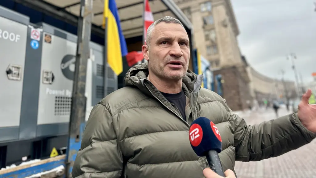 En person i en grå dunjacka håller ett mikrofonmärke från en nyhetskanal framför sig. I bakgrunden syns ukrainska och andra flaggor samt en gata i en stadsmiljö.