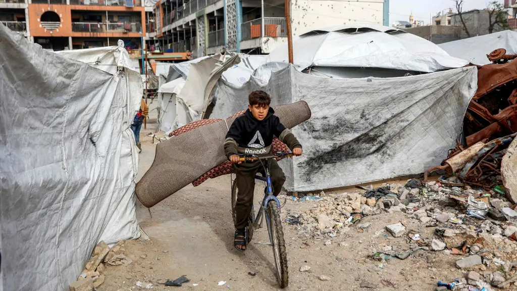En pojke cyklar genom ett område med tält och skräp i Gaza. Tälten är provisoriska och omgivningen präglas av förstörelse. Himlen är mulen och byggnader syns i bakgrunden.