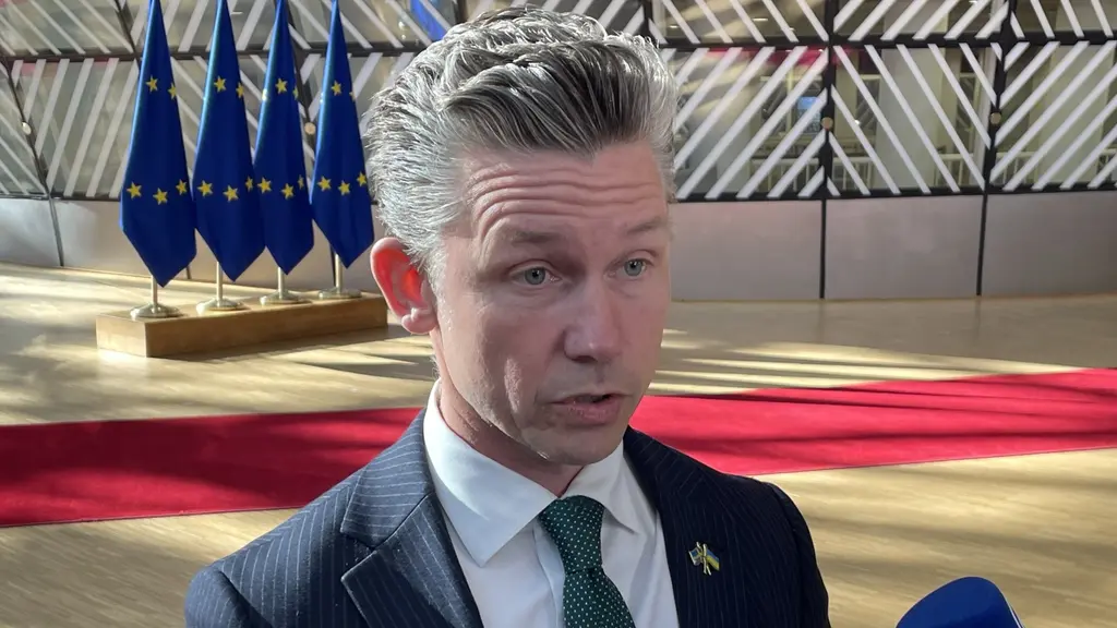 En man i kostym med en grön slips intervjuas av journalister som håller fram mikrofoner. I bakgrunden syns EU-flaggor och en röd matta i en byggnad med glasväggar.